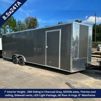 Charcoal gray enclosed cargo trailer with dual axles, side entry door, and diamond-plate front, on a gravel lot under a blue sky.